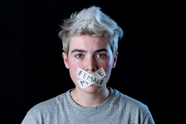 A trans male teenager with “Male”/ “Female” stickers taped over their mouth looks straight into the camera.