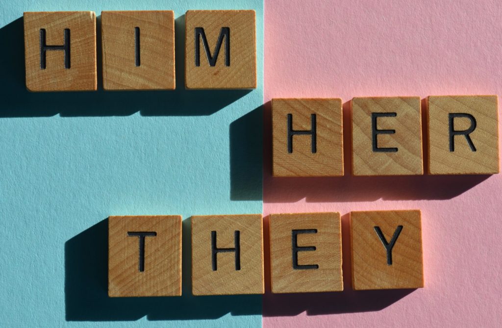A blue and pink background with wooden blocks spelling “Him,” “Her,” and “They.” 