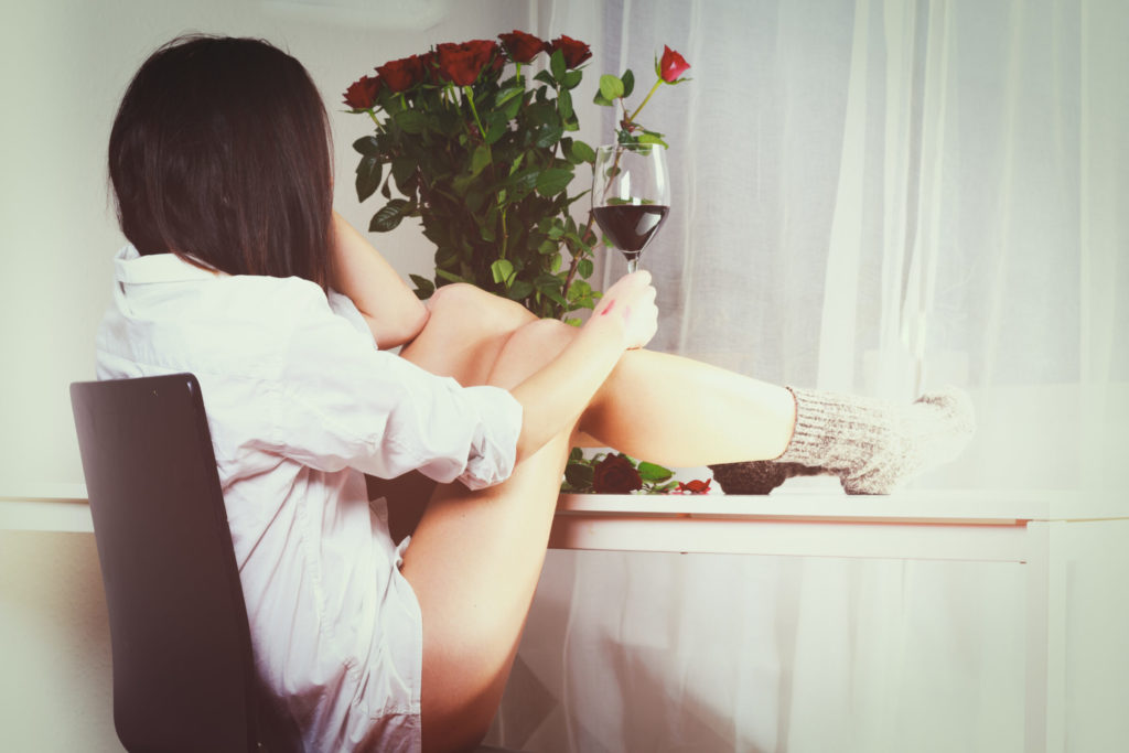 A woman facing away from the camera with her legs up on a table drinking red wine and looking at a bouquet of red roses.
